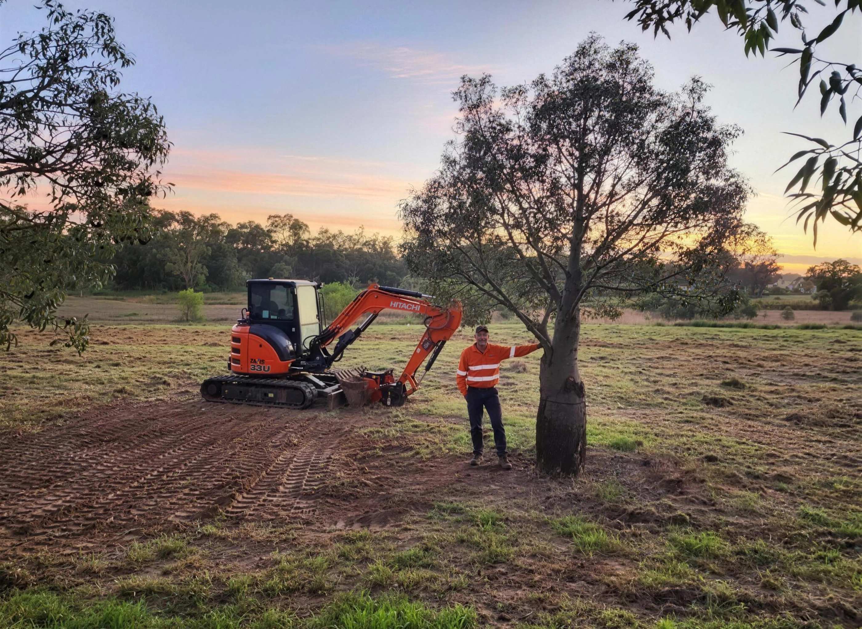 Russell Clark resting a hand on a bottle tree at sunset, a small excavator parked behind him